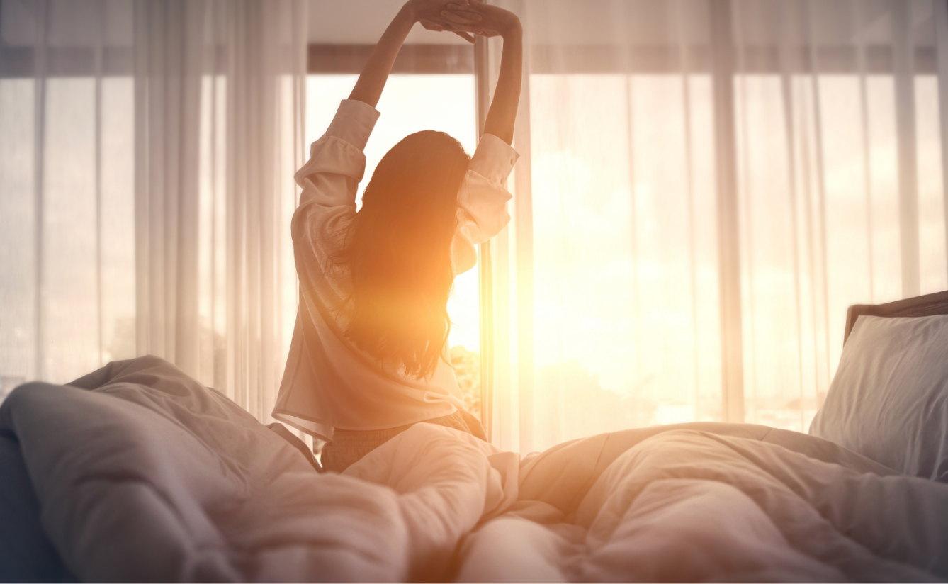 Eine Person sitzt im Bett, streckt sich nach dem Aufwachen mit dem Rücken zum Fenster in der hellen Morgensonne.