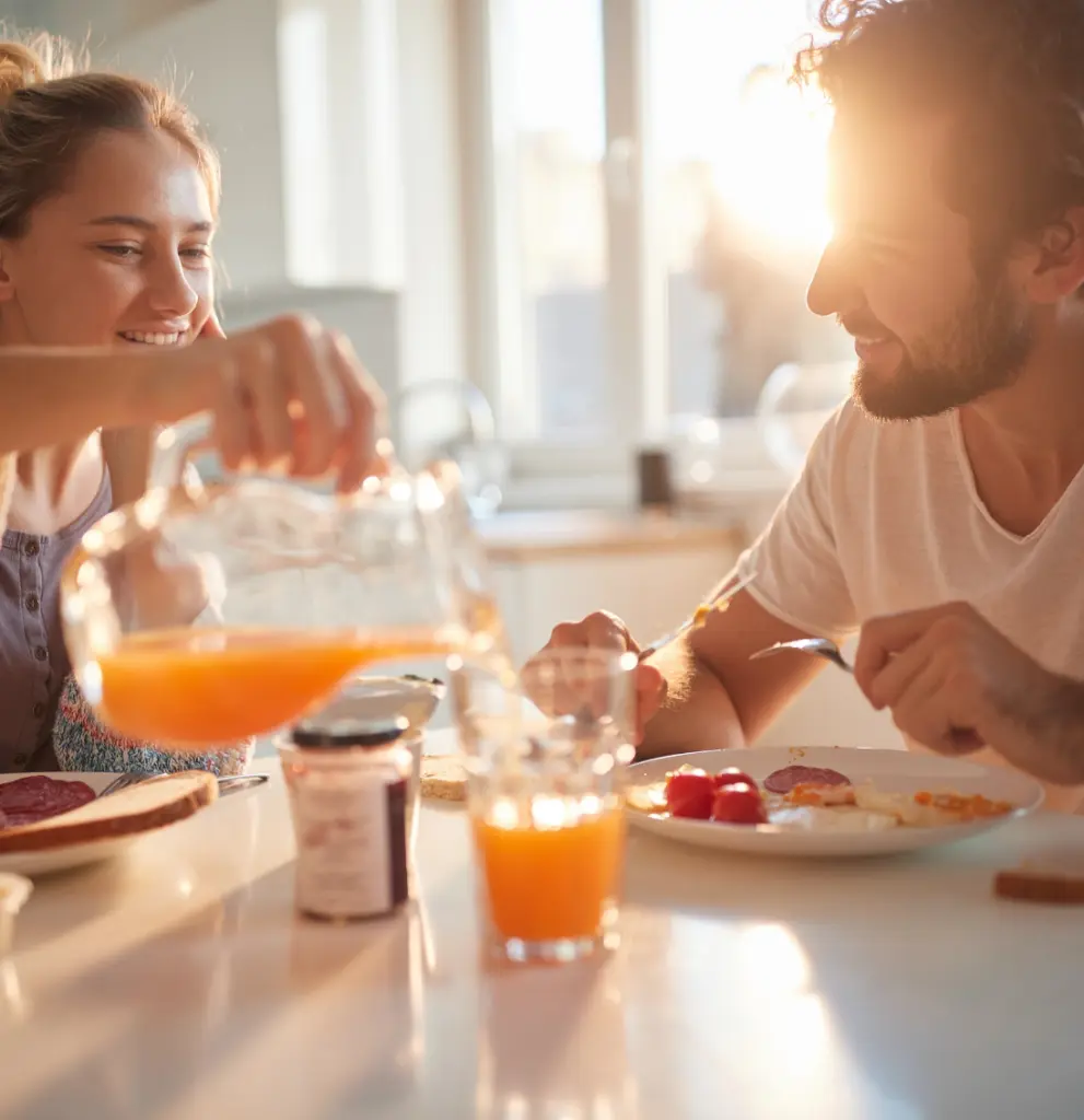 Ein Paar frühstückt in einer sonnendurchfluteten Küche, die Frau gießt Orangensaft ein.