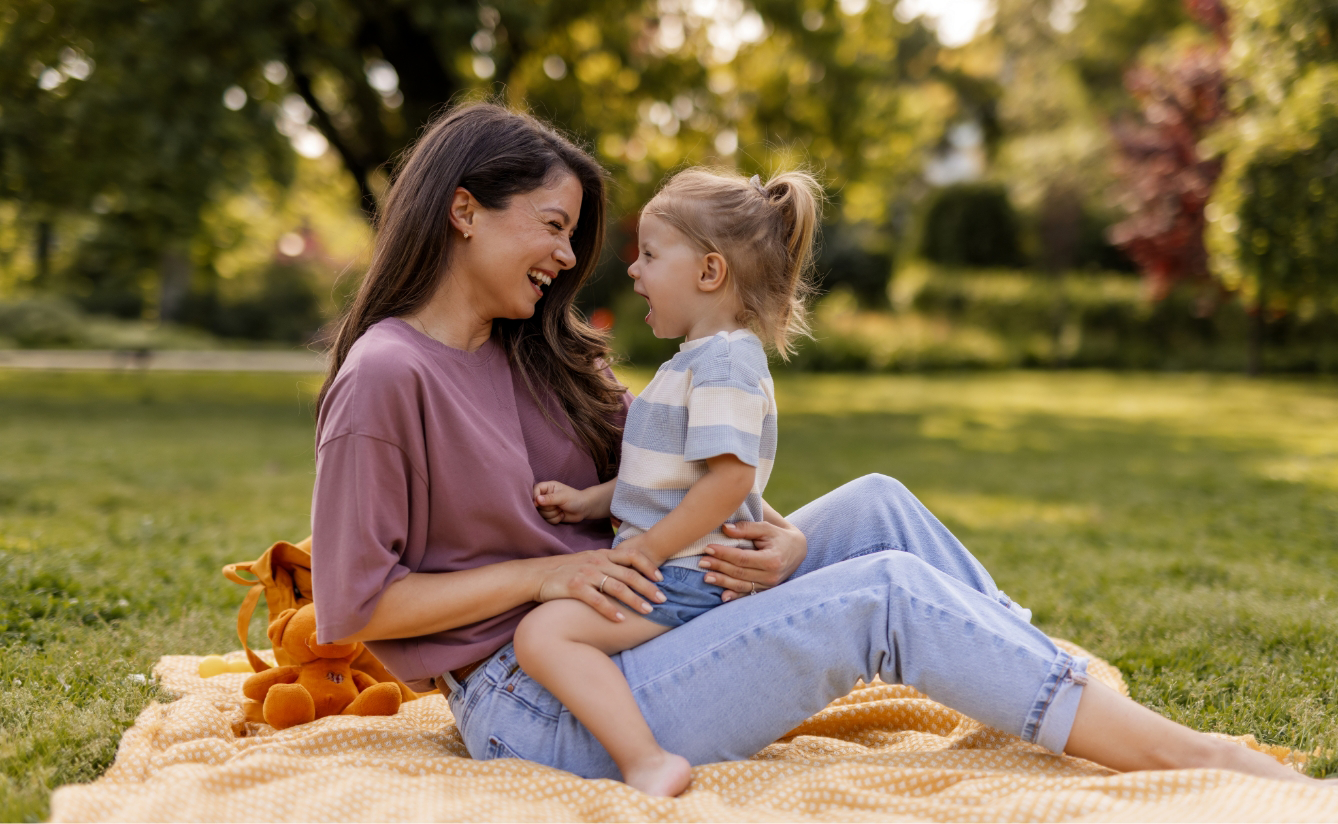 Eine Frau sitzt lächelnd mit einem Kleinkind auf dem Schoß auf einer gelben Decke im Gras in einem Park.